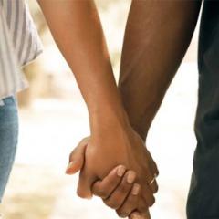 Close-up of a man and woman holding hands, focusing on their intertwined fingers, with partial views of their casual attire in a softly blurred background.